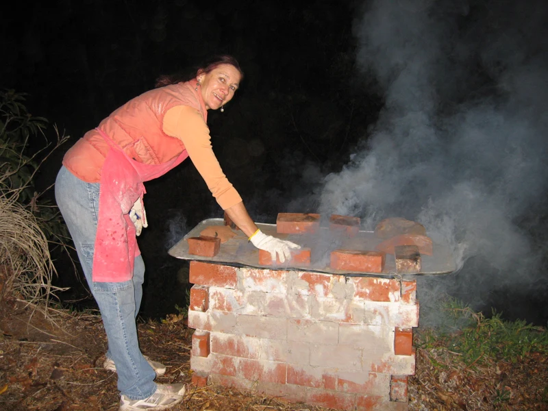 Francine tending an outdoor brick firing pit as smoke rises.